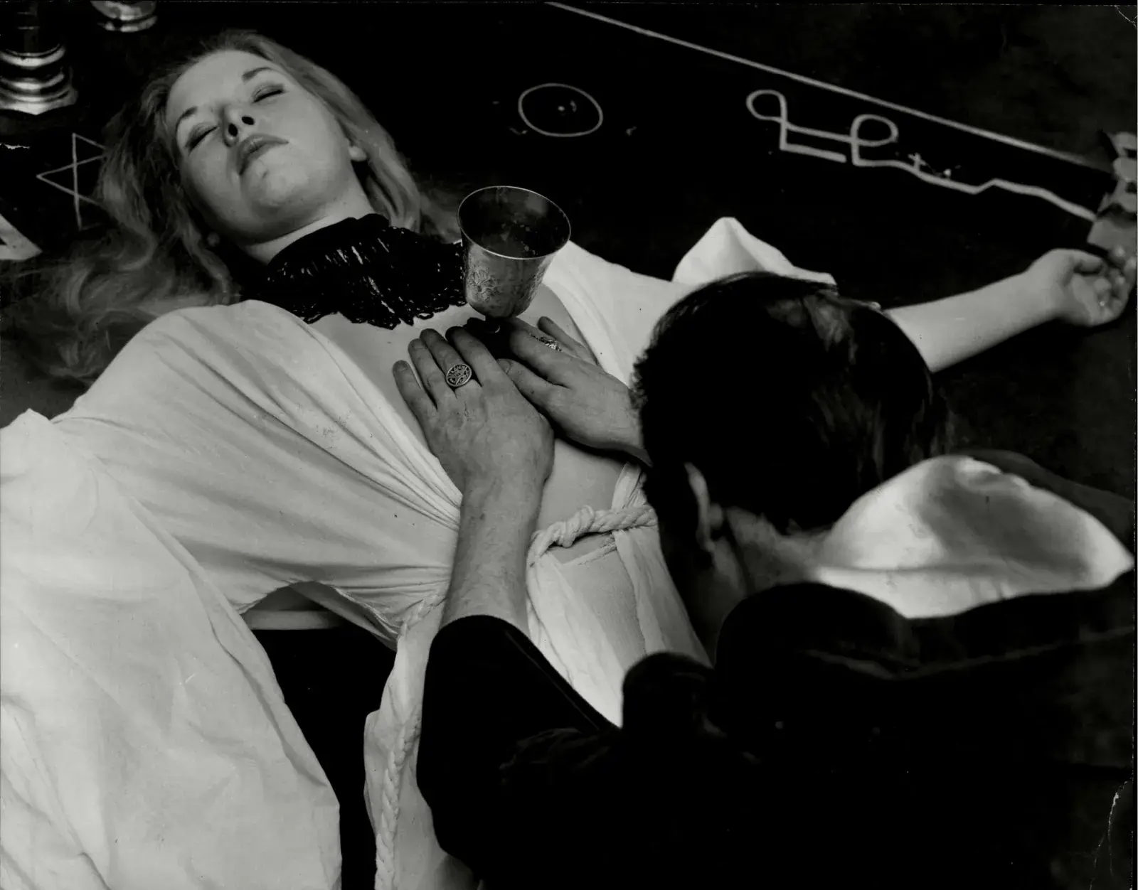 A black-and-white photograph of a ritual: a woman in white robes lies with her eyes closed and a chalice resting on her chest, while a dark-haired man in dark clothes bends over her, symbols drawn on the floor around them.