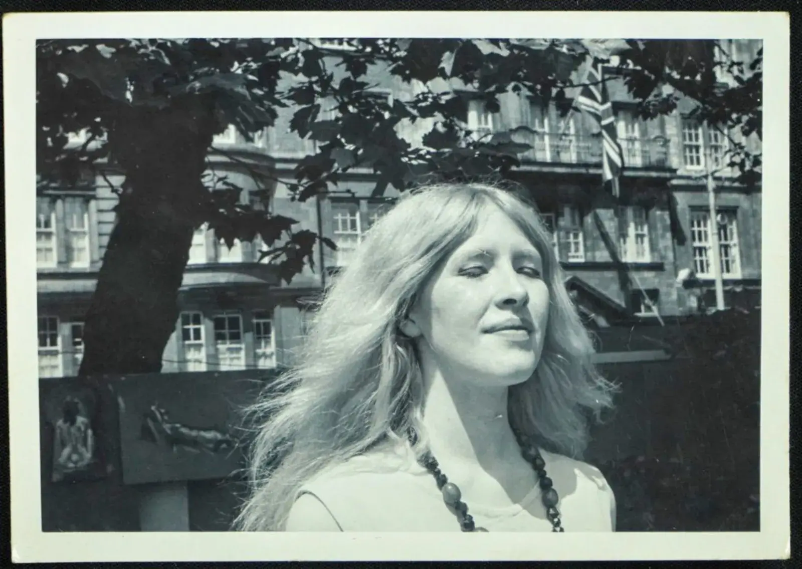 A black-and-white photograph of Maxine Sanders as a young woman, her fair hair loose and her face lifted to the sun, with a tree and a tall building behind her.